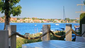 A wooden table with rope barriers, overlooking a body of water with boats and houses in the background. - Hotel Amax (Mikolajki)