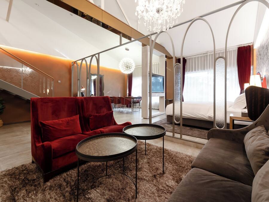 A modern hotel room with a red sofa, a brown sofa, two small round tables, a chandelier, and a large bed with a TV in the background.