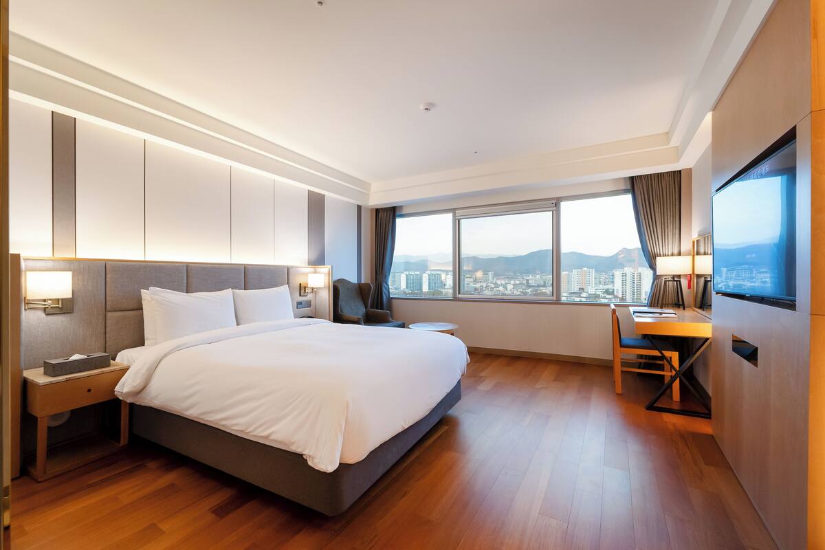A modern hotel room with a large bed, a desk, and a view of the cityscape.
