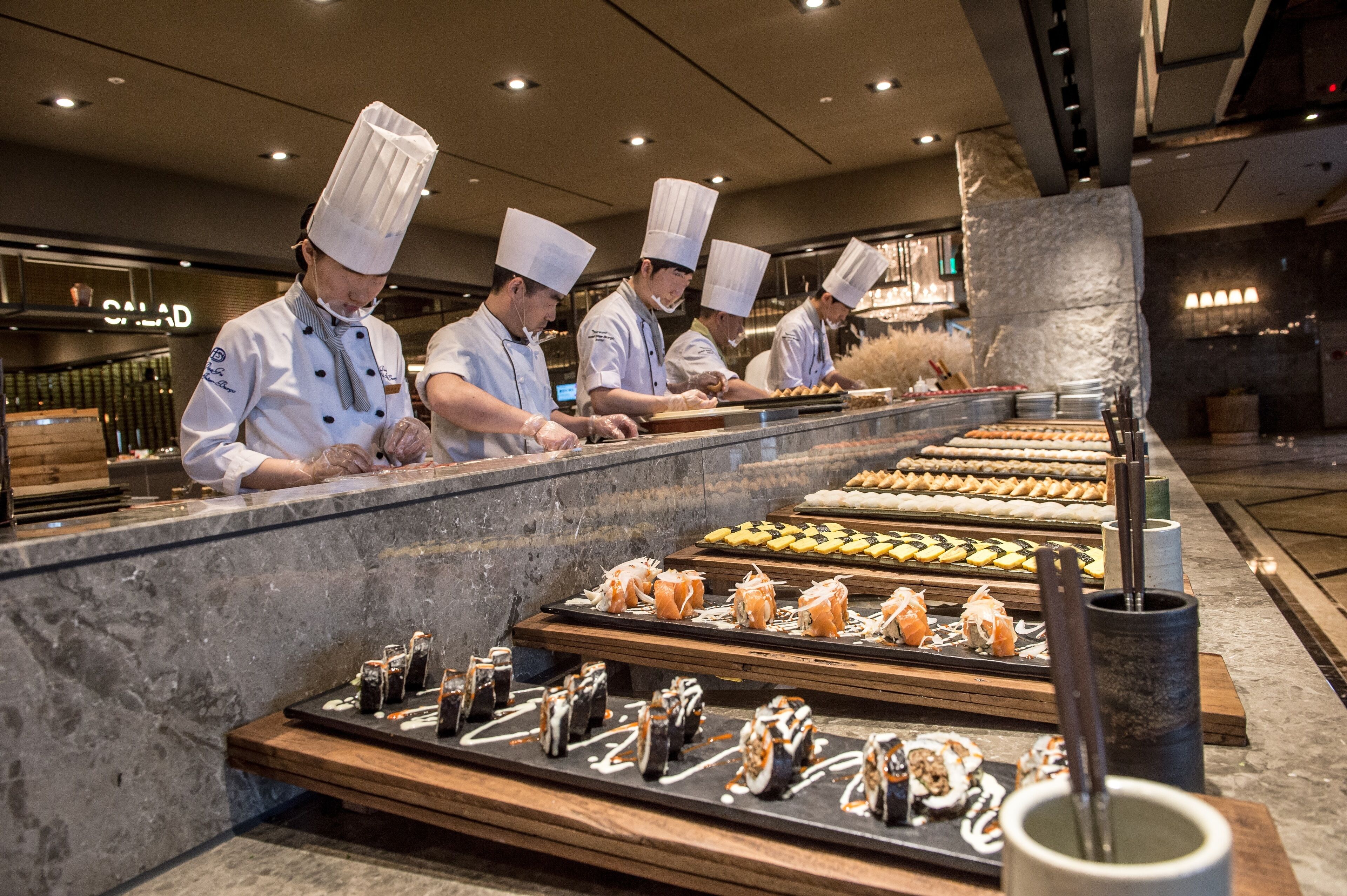 A sushi bar with chefs preparing dishes, a variety of sushi displayed on trays, and a stone pillar in the background.