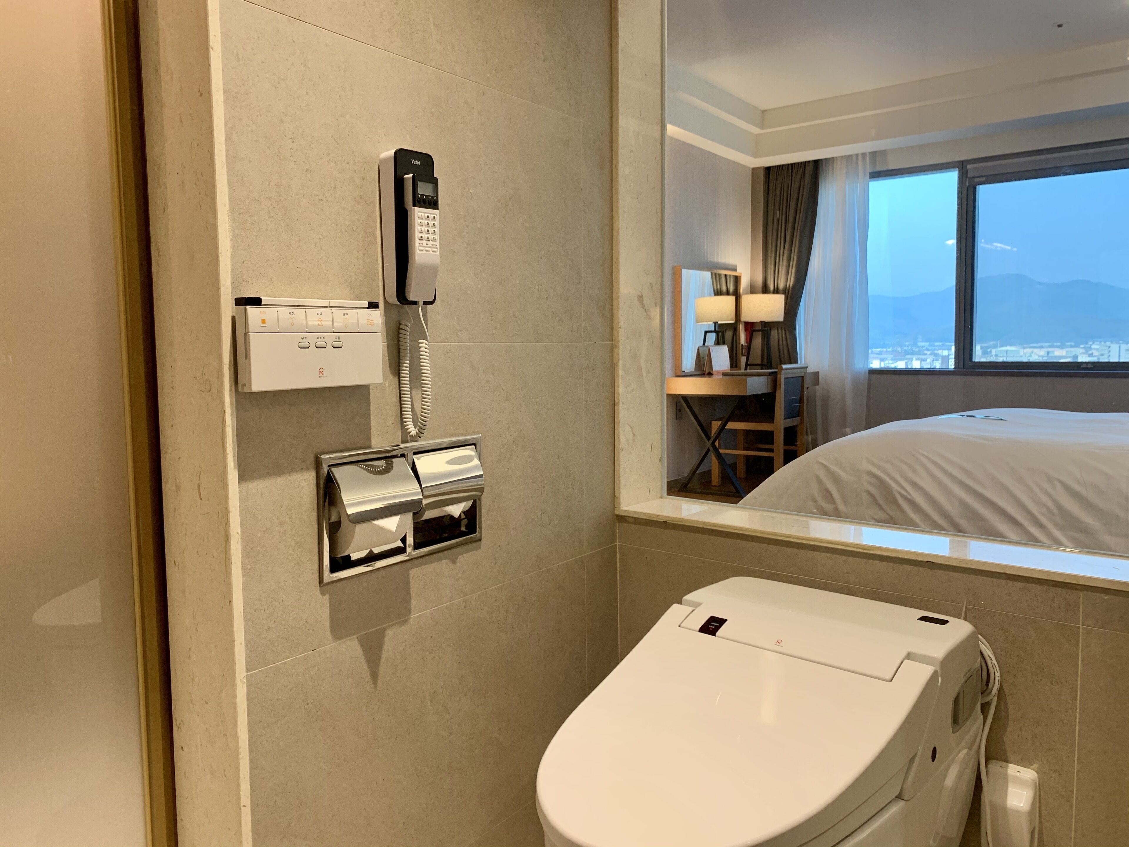 A hotel bathroom with a toilet, a wall-mounted telephone, and a window with a city view.