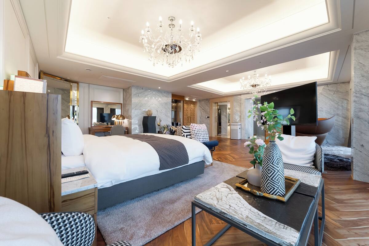 A modern hotel room with a large bed, a desk, a TV, and a chandelier.