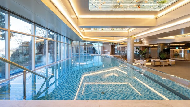 A modern indoor swimming pool with glass railings, a geometric tile pattern, and a ceiling with a skylight.