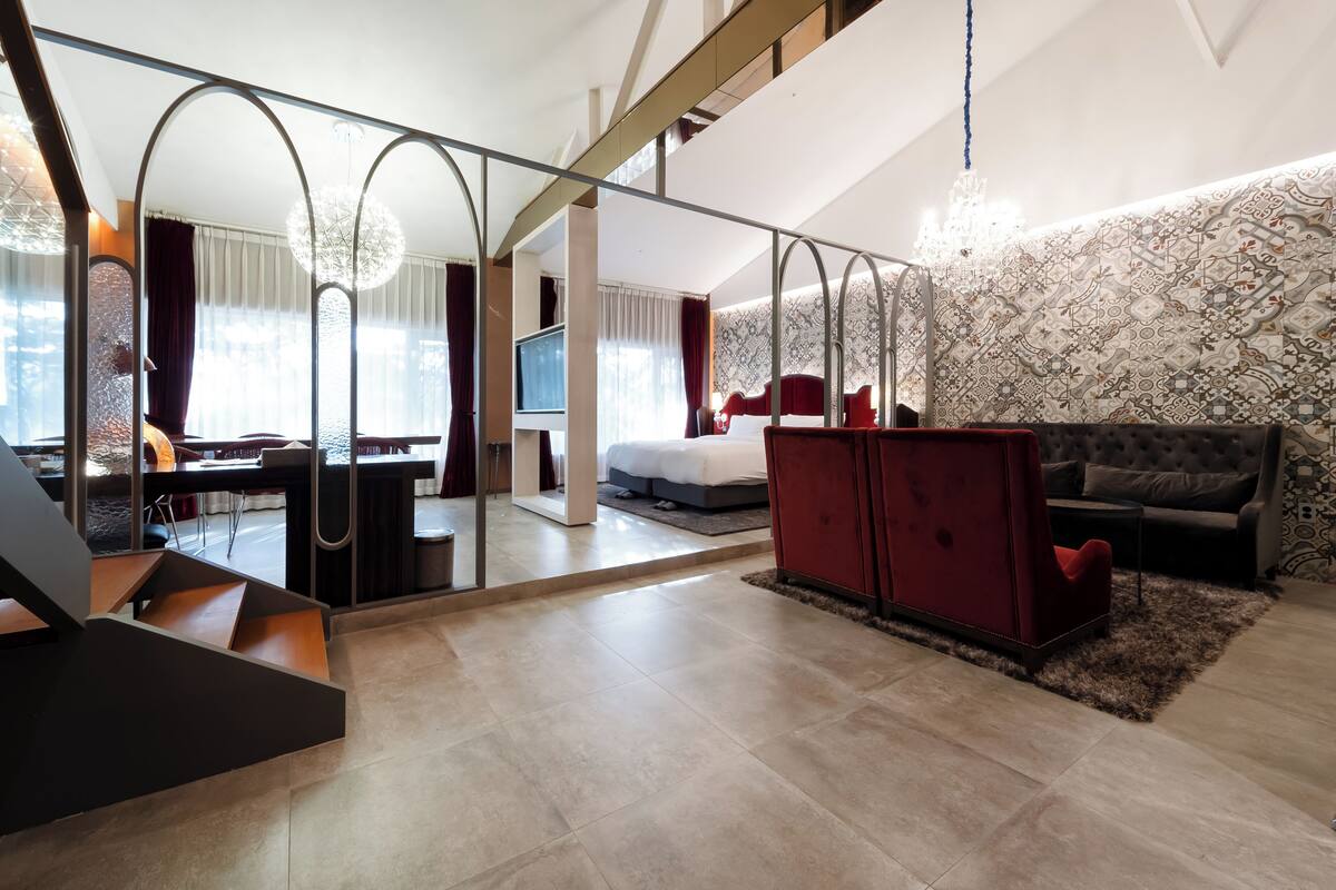 A modern hotel room with a large bed, a red sofa, a TV, and a dining area with a table and chairs.