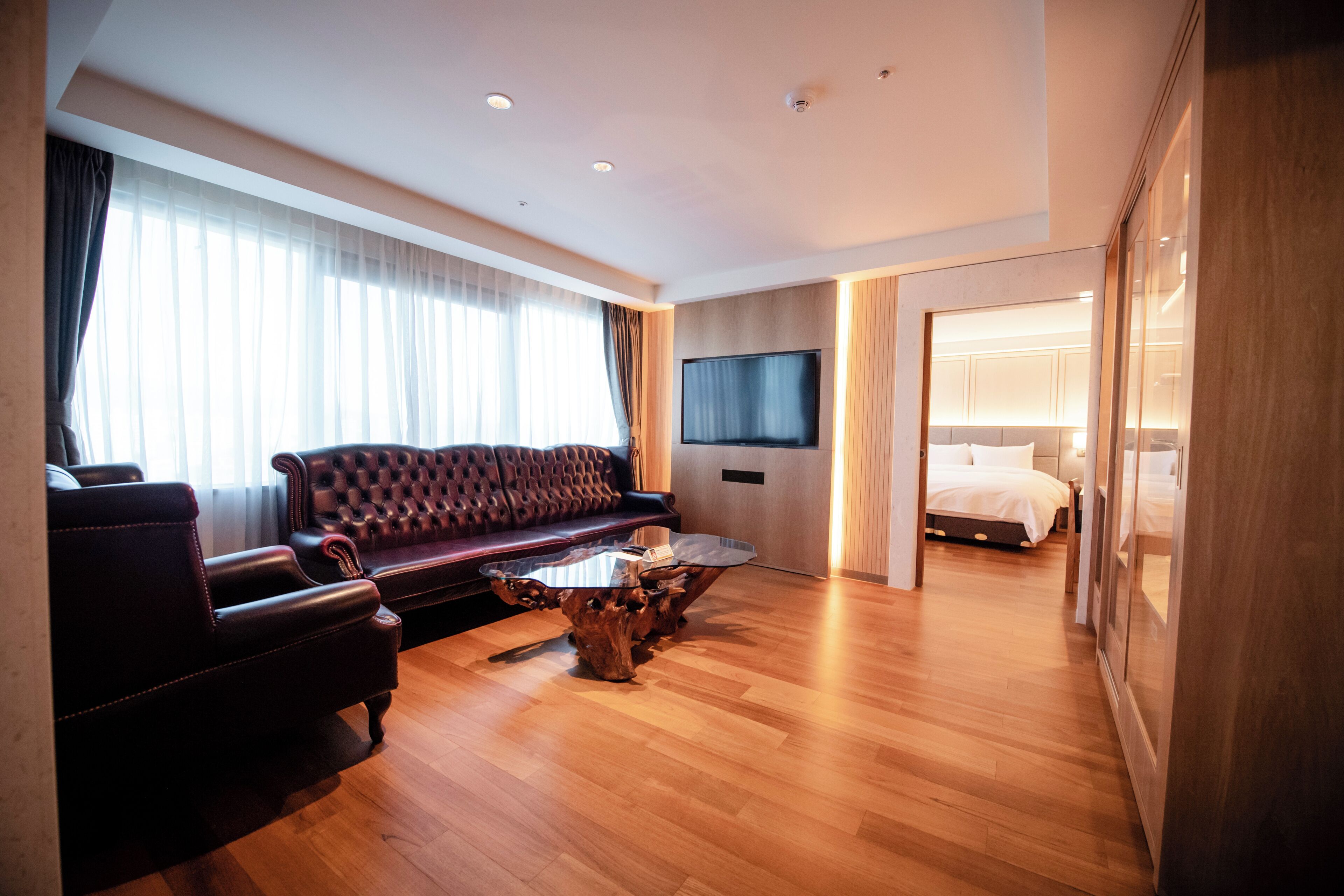 Une chambre d’hôtel moderne avec un canapé en cuir, une table basse en bois, un téléviseur à écran plat et un lit recouvert de draps blancs.