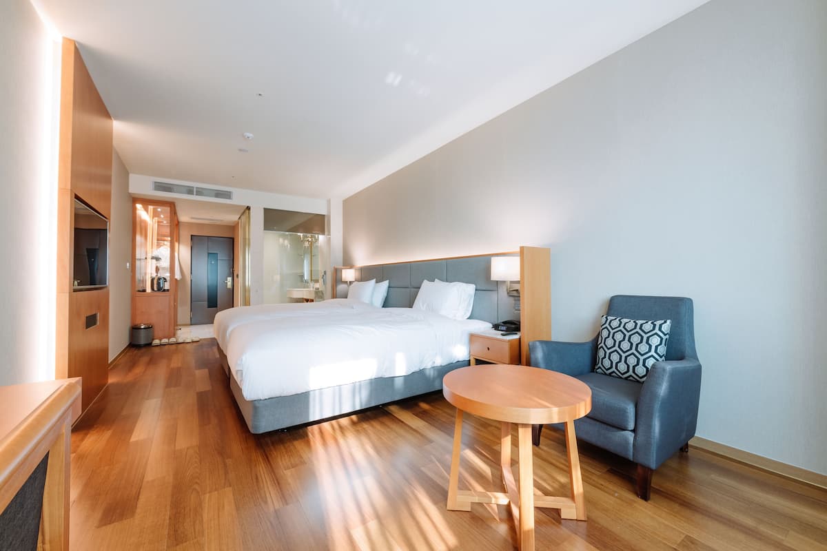 A hotel room with a large bed, a small round table, a blue armchair, a nightstand, and a mirror on the wall.