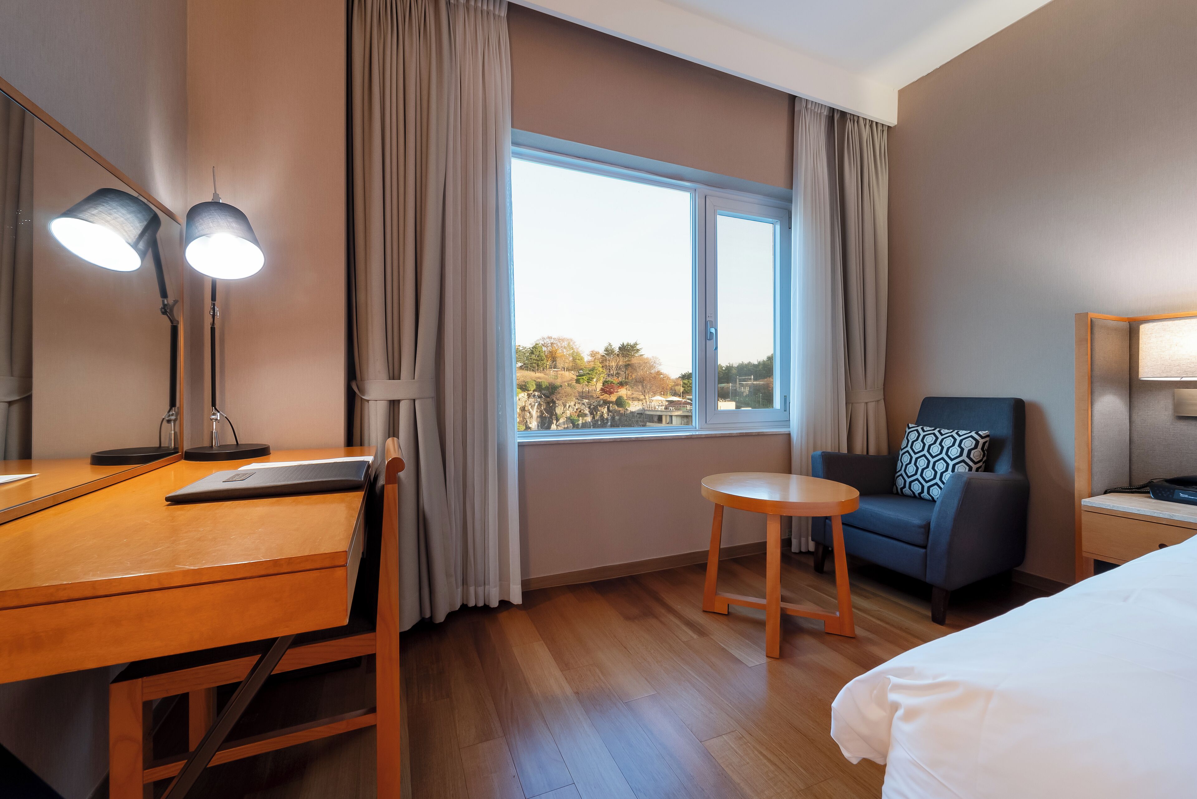 Habitación de hotel con un escritorio de madera, dos lámparas, una silla, una mesita redonda pequeña, una cama y un ventanal grande con cortinas.