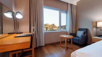 Habitación de hotel con un escritorio de madera, dos lámparas, una silla, una mesita redonda pequeña, una cama y un ventanal grande con cortinas.