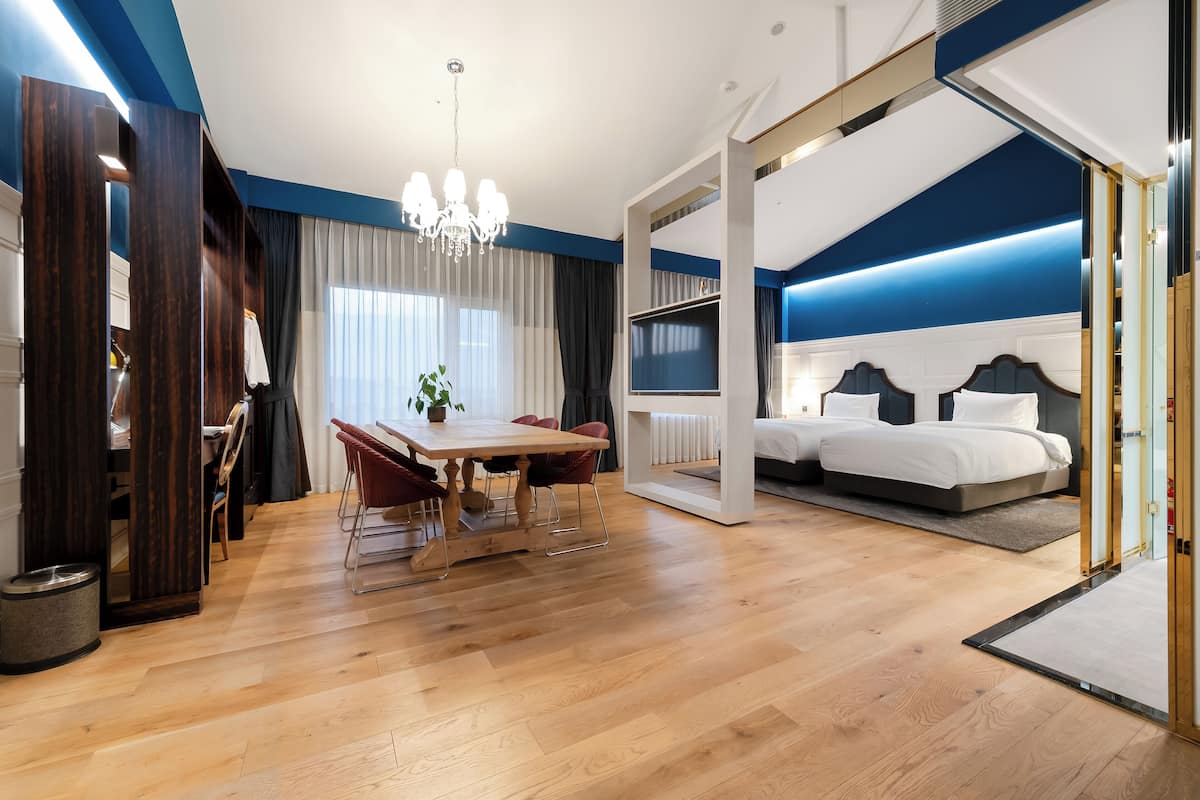 A modern hotel room with a wooden dining table, a chandelier, a bed with a headboard, and a TV.