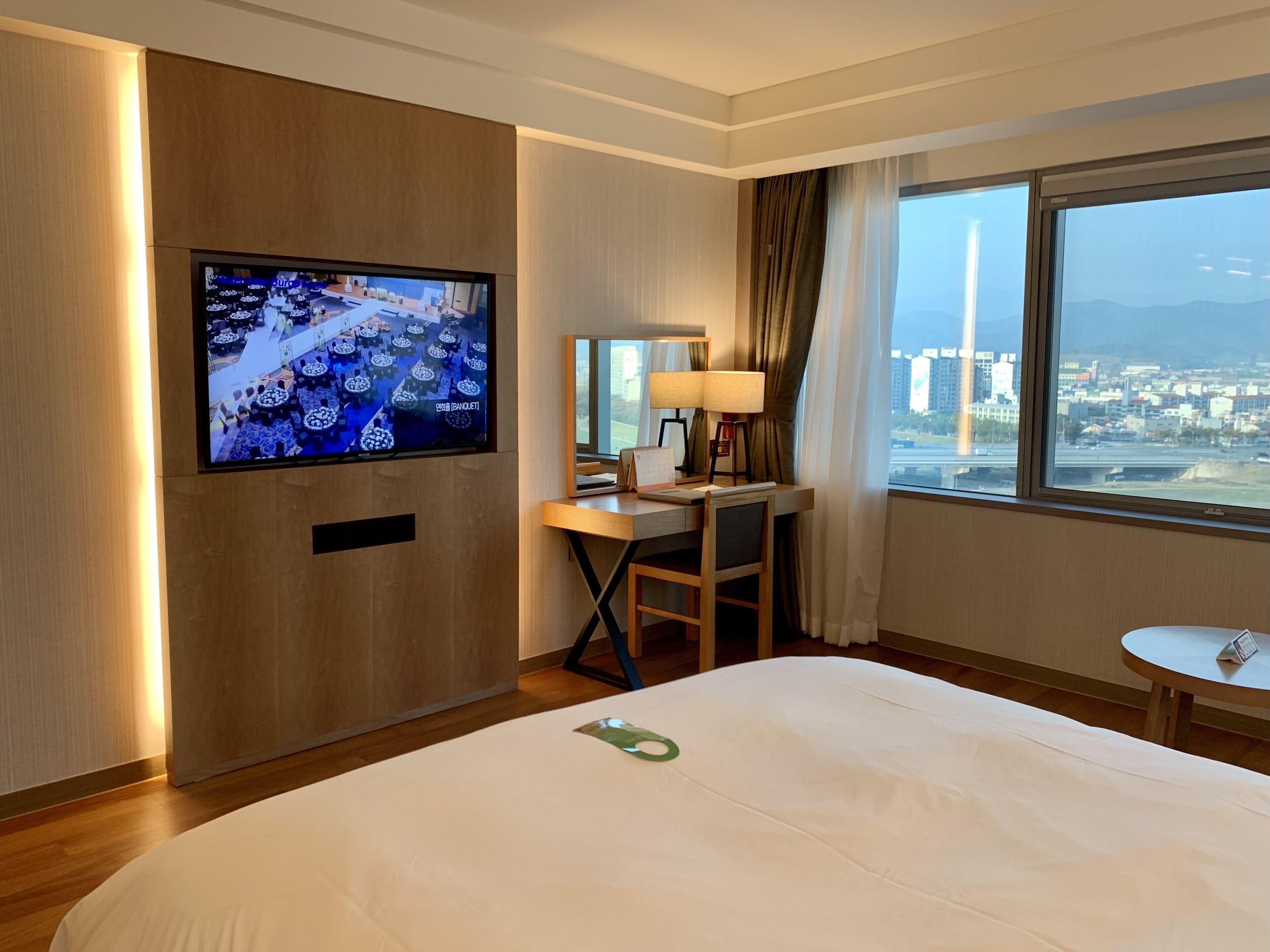 Une chambre d’hôtel comprenant un lit, un bureau avec une chaise, un téléviseur fixé au mur, une fenêtre donnant sur la ville et une lampe.