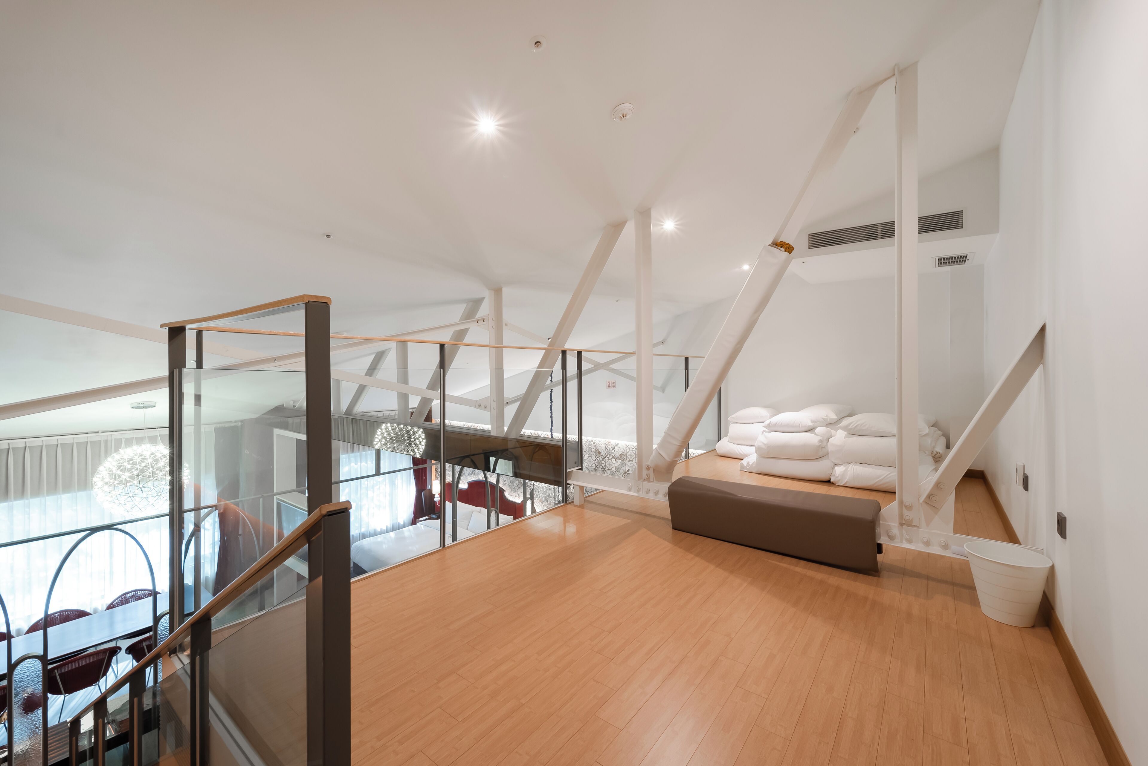 Un'ampia camera da letto mansardata con soffitto alto, balcone con ringhiera in vetro e un'accogliente area salotto con cuscini.