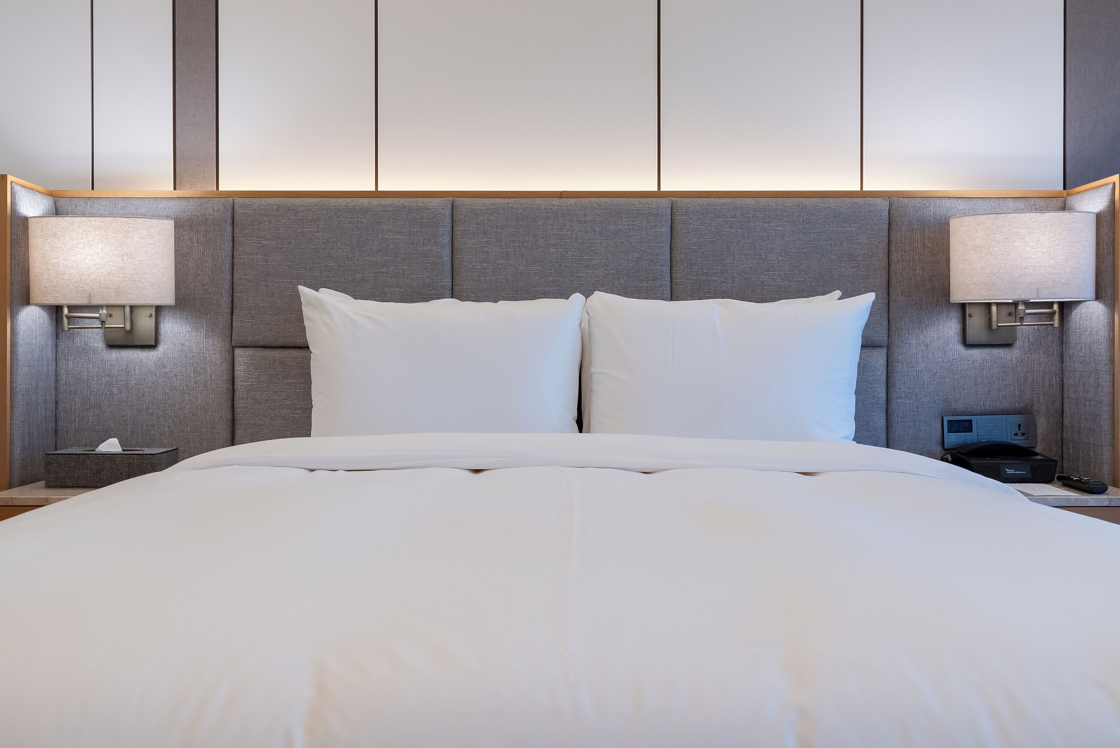 Una cama bien hecha con sábanas blancas, dos lámparas de noche y una mesita de noche con una caja de pañuelos.