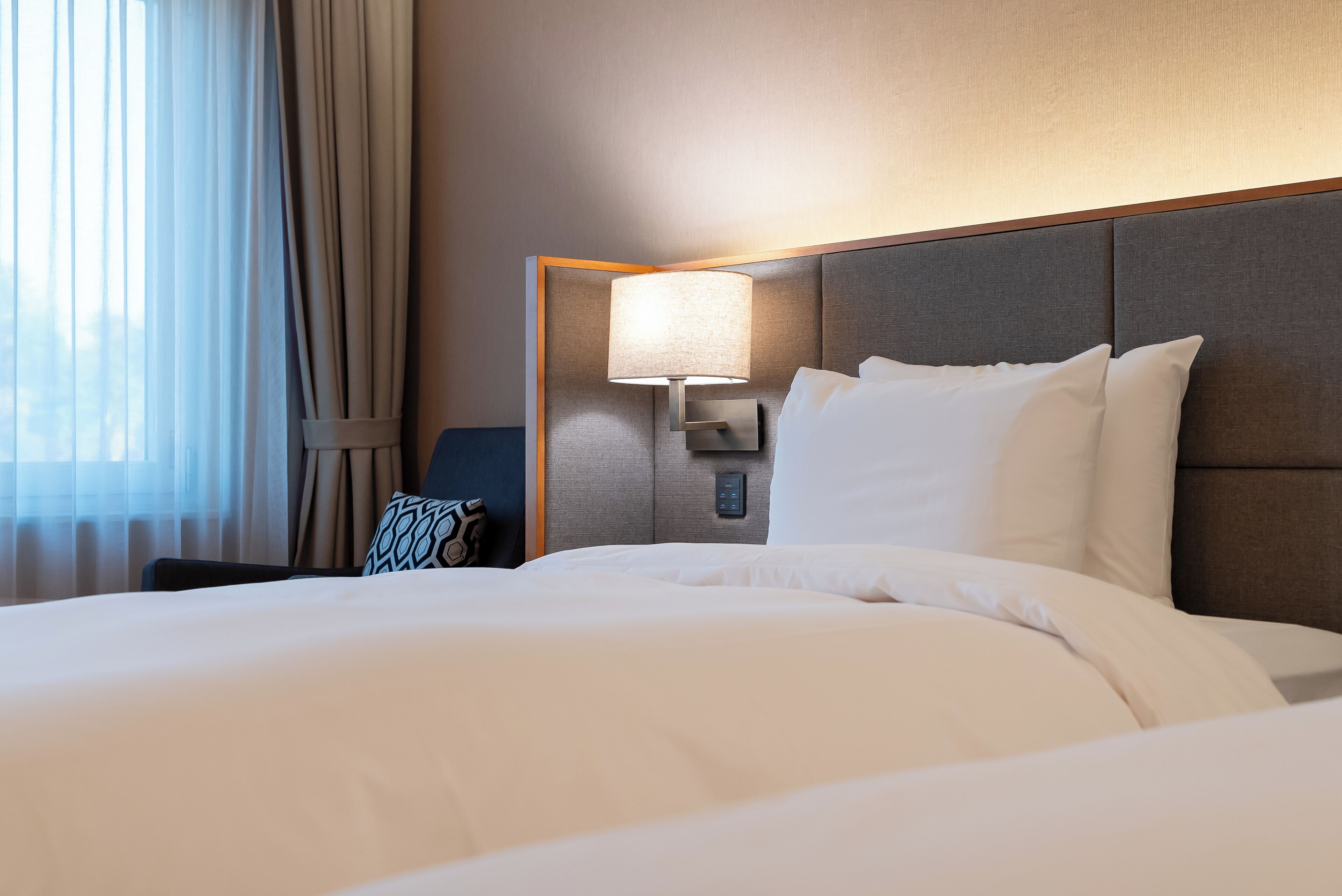 Una habitación de hotel con una cama grande, una lámpara de noche y vistas al exterior a través de cortinas traslúcidas.