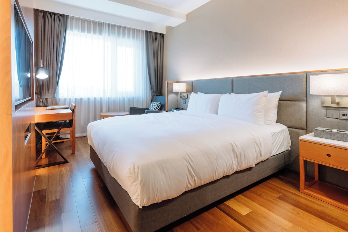 A hotel room with a large bed, a desk, a chair, a bedside table, and a lamp.