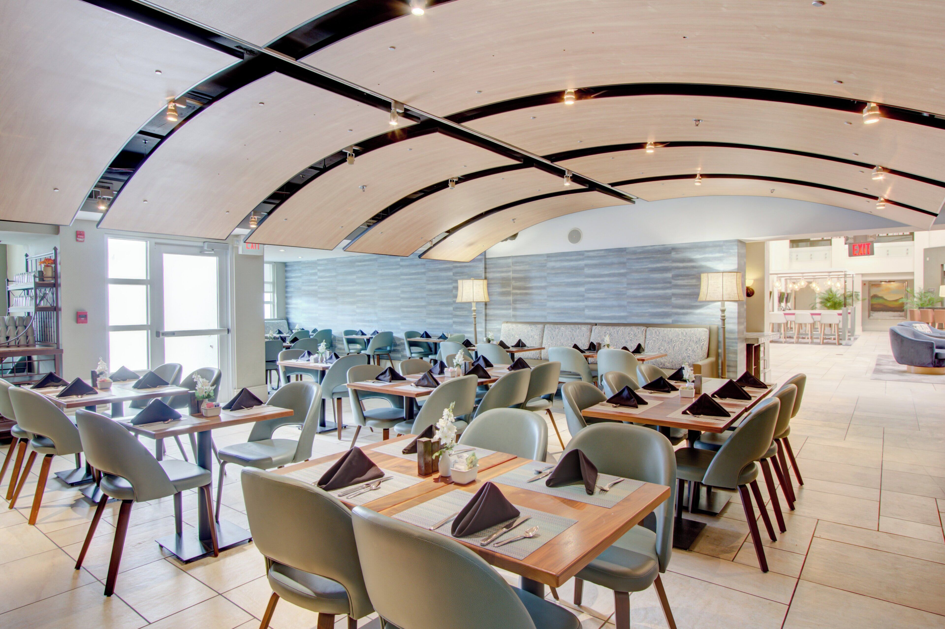A spacious dining area with multiple tables and chairs, a high ceiling with a unique curved design, and a view of a restaurant in the background.