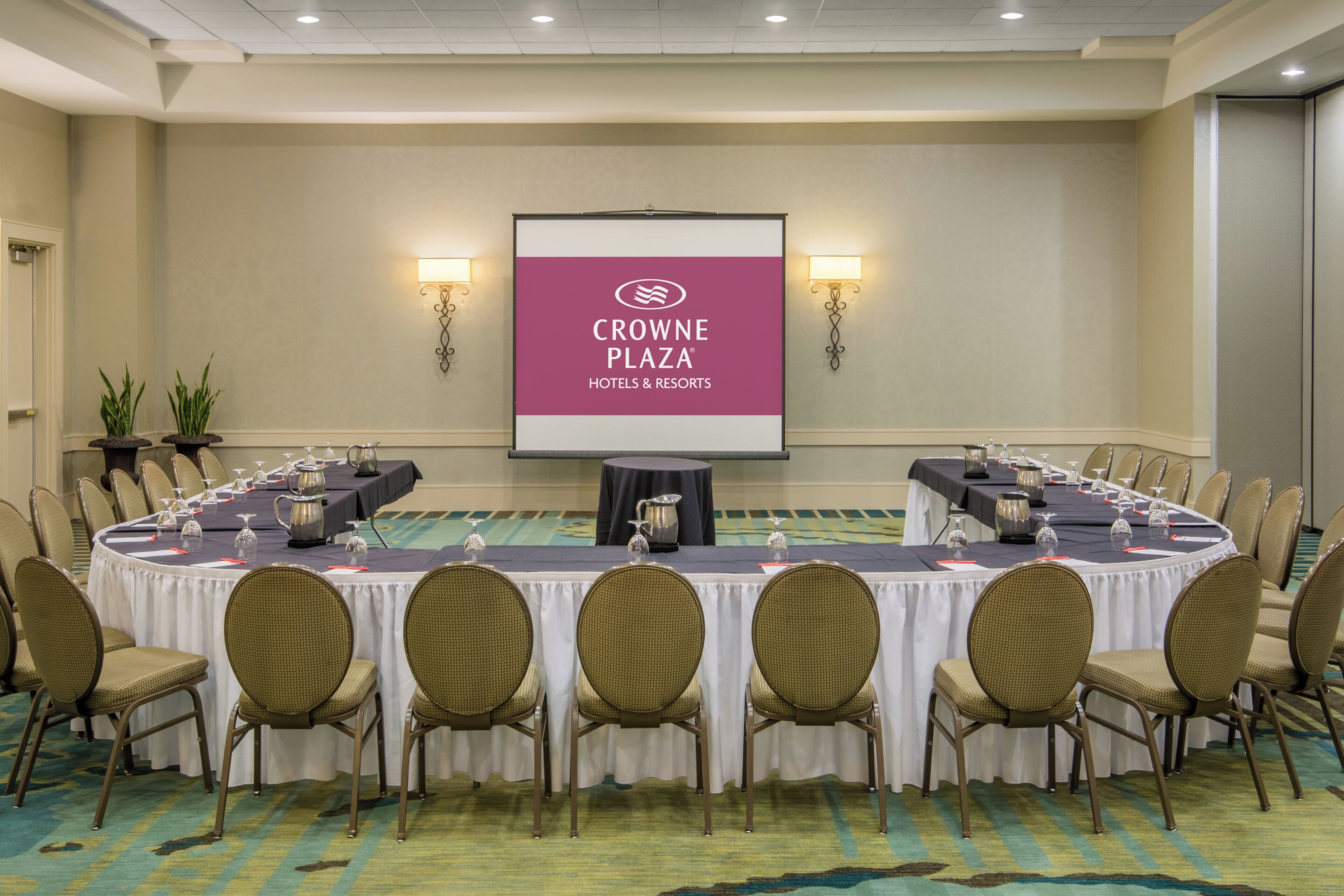 Ett konferensrum med ett långt bord, stolar och en skärm som visar varumärket förkedjan «CROWNE PLAZA HOTELS & RESORTS».