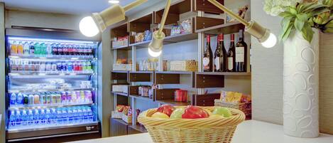 Zona comercial bien iluminada con refrigerador de bebidas, estantes llenos de bocadillos y botellas de vino, y una cesta de mimbre con frutas.