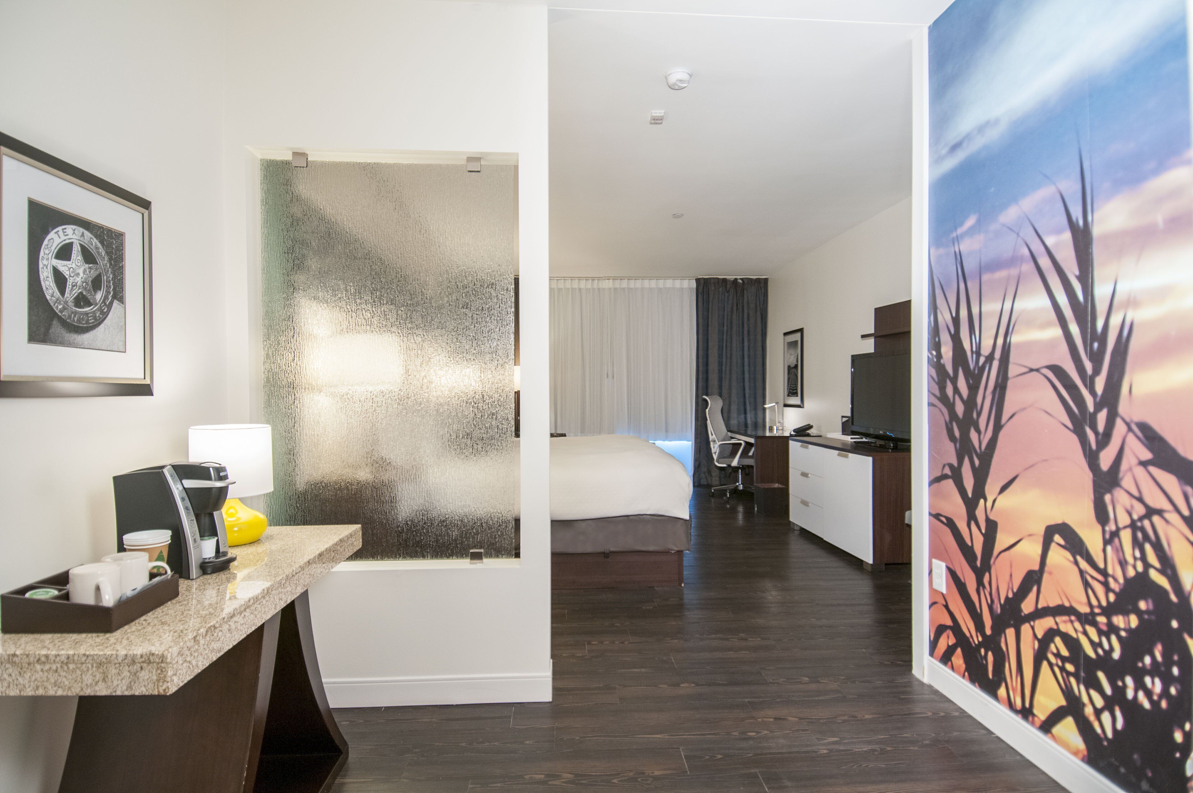 A hotel room with a bed, a desk with a coffee maker, a chair, and a painting of tall grass.