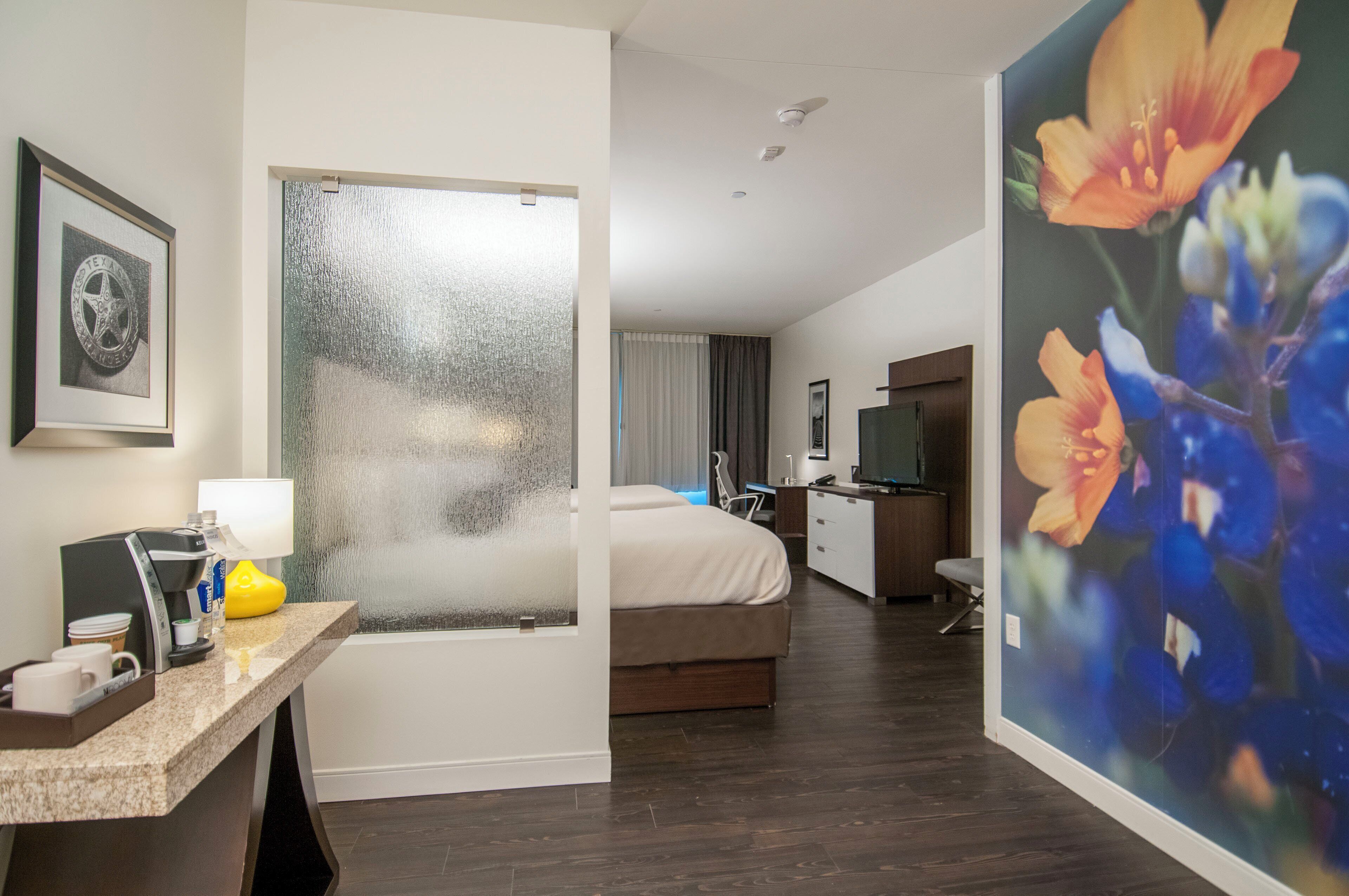 A hotel room with a bed, a desk with a coffee maker, a TV, and a painting on the wall.