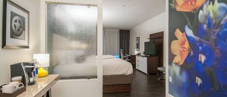 A hotel room with a bed, a desk with a coffee maker, a TV, and a painting on the wall.