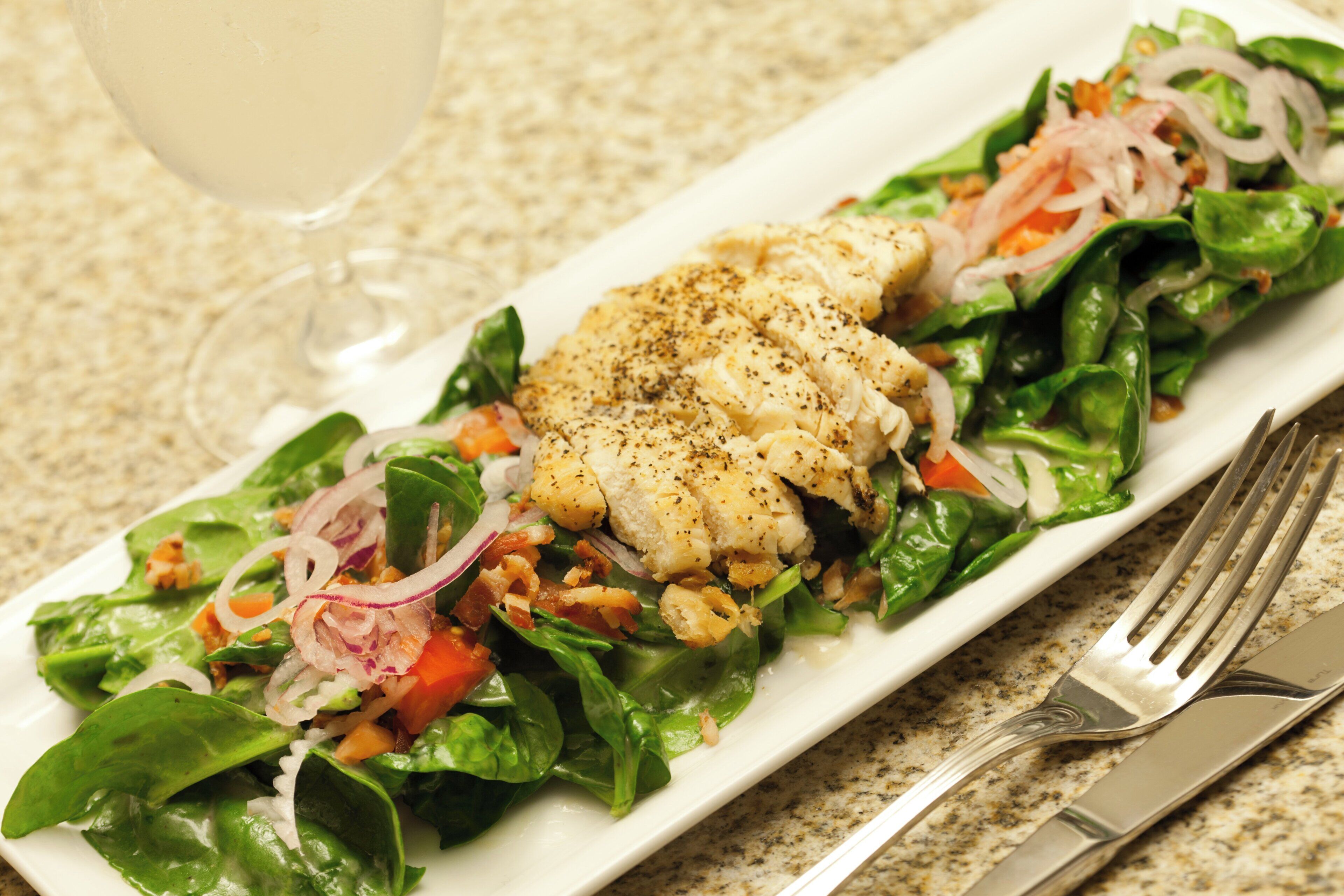 A rectangular plate with a salad containing greens, tomatoes, onions, and a piece of grilled fish, accompanied by a fork and knife on the right.