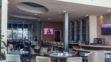 Eine moderne Bar mit Sitzgelegenheiten, einem Fernsehbildschirm und Blick auf die Stadt durch große Fenster.