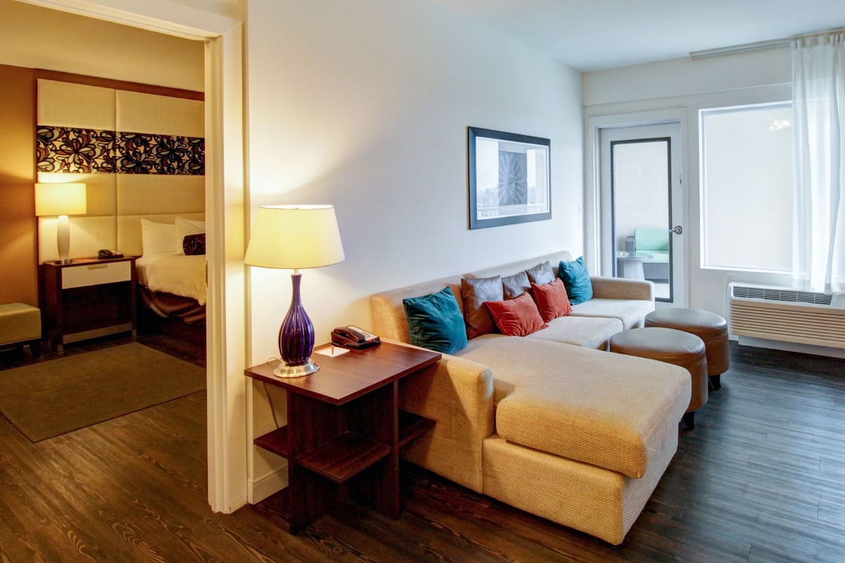A hotel room with a sofa, a bedside table with a lamp, a chair, a bed, and a large window with curtains.