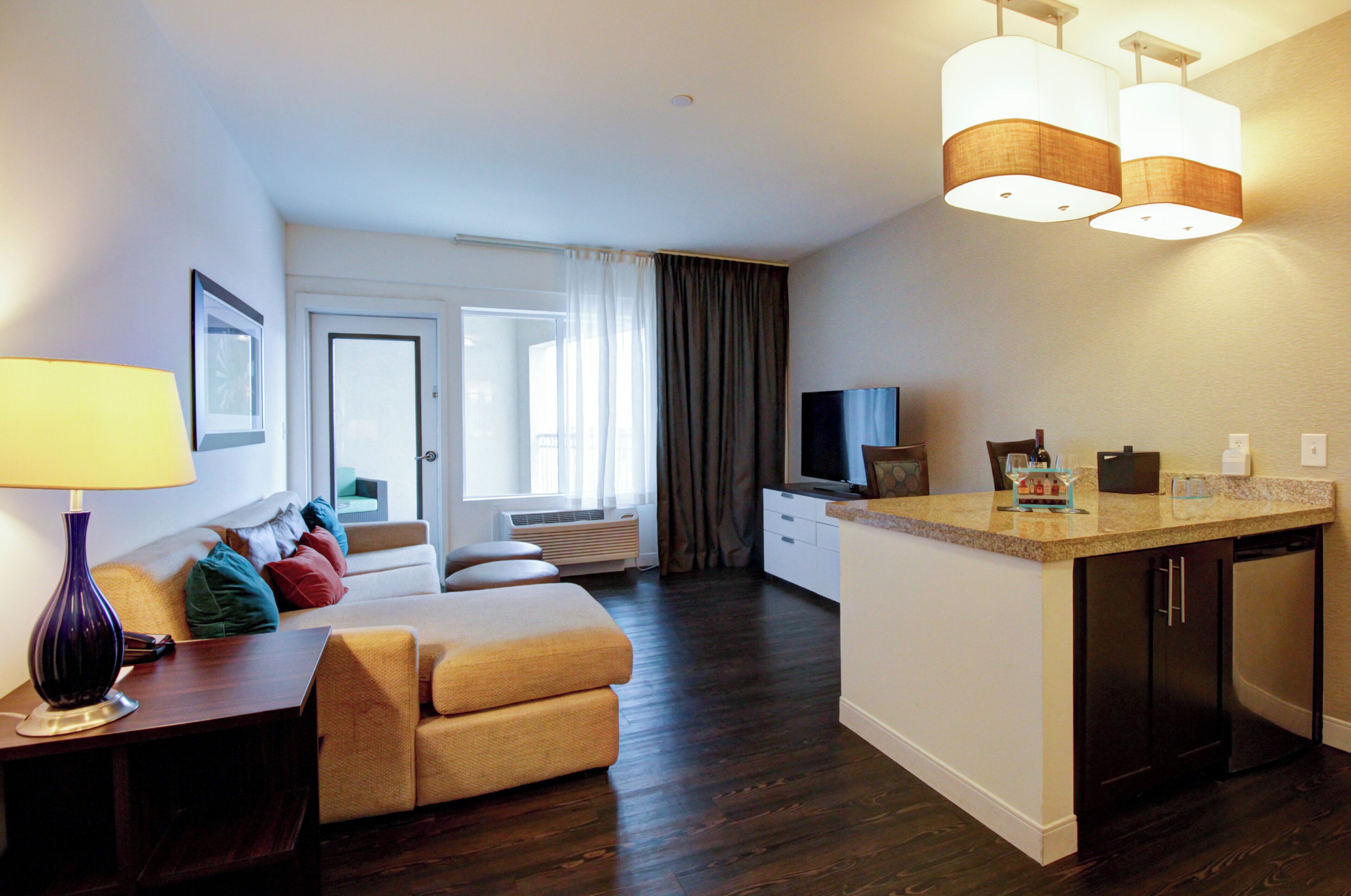 A modern hotel room with a sofa, a small dining table, a TV, and a kitchen area with a countertop and cabinets.