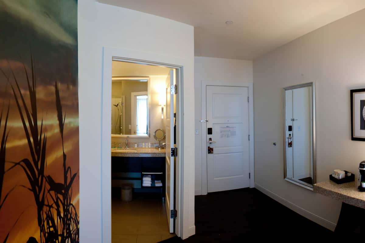 A hotel hallway with a bathroom visible through an open door, featuring a sink, mirror, and towel rack.