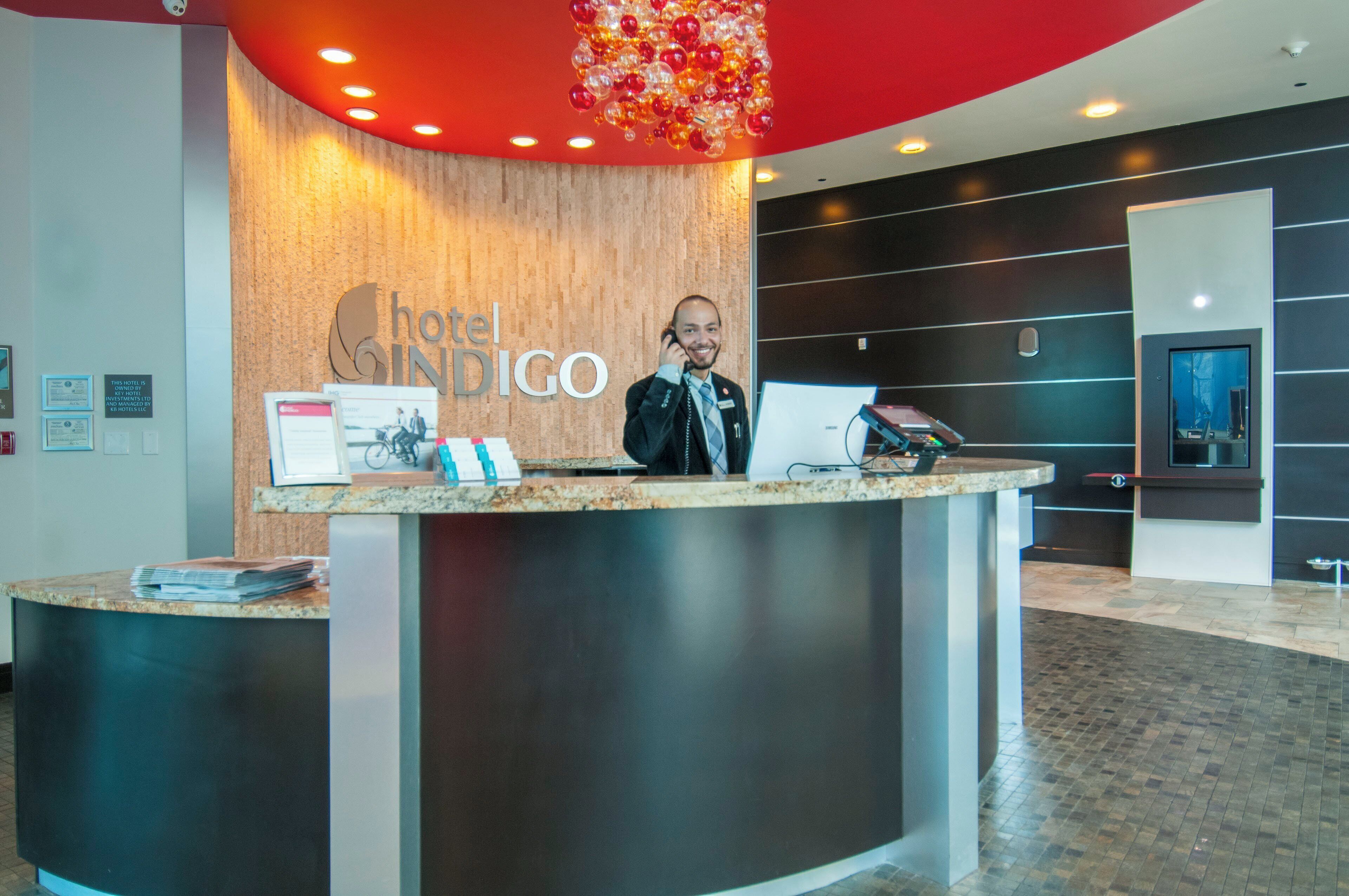 Un comptoir d’accueil d’hôtel au design moderne, une plaque nominative indiquant « Hotel Indigo », et un écran numérique affichant des informations.