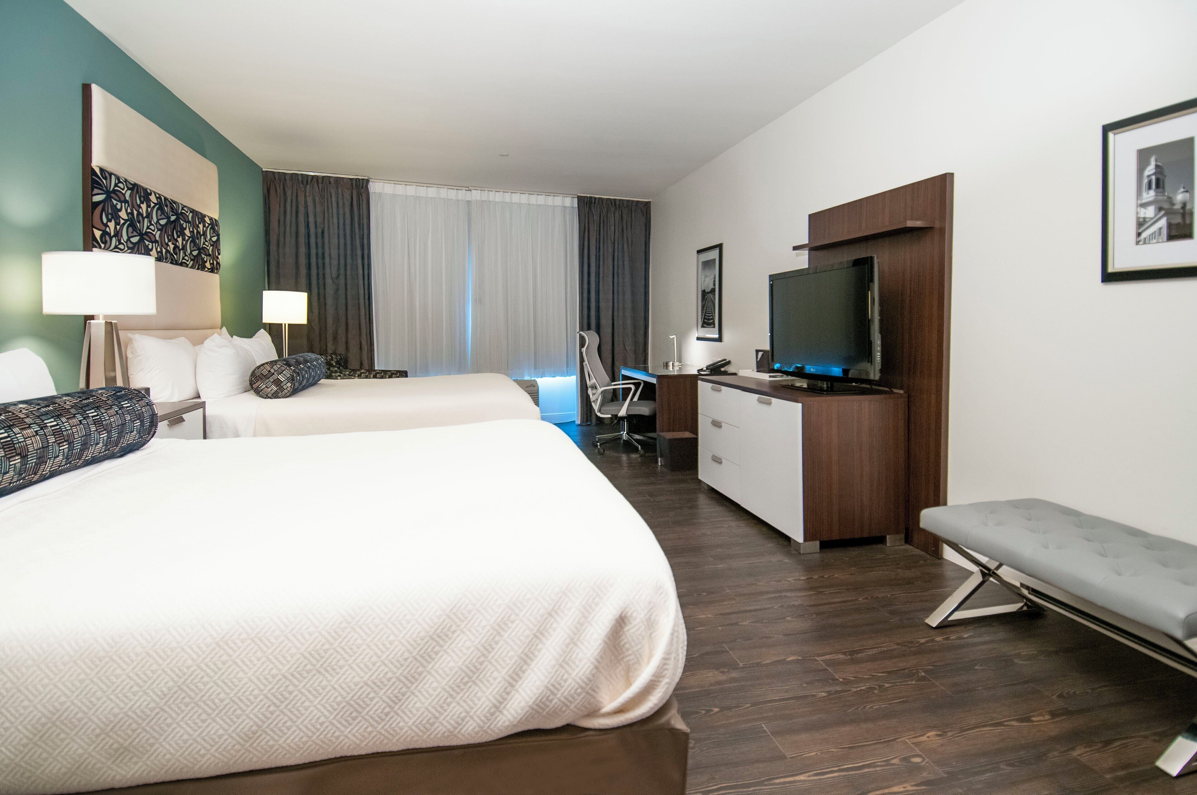 A hotel room with two beds, a desk with a TV, a bench, and a large window with curtains.