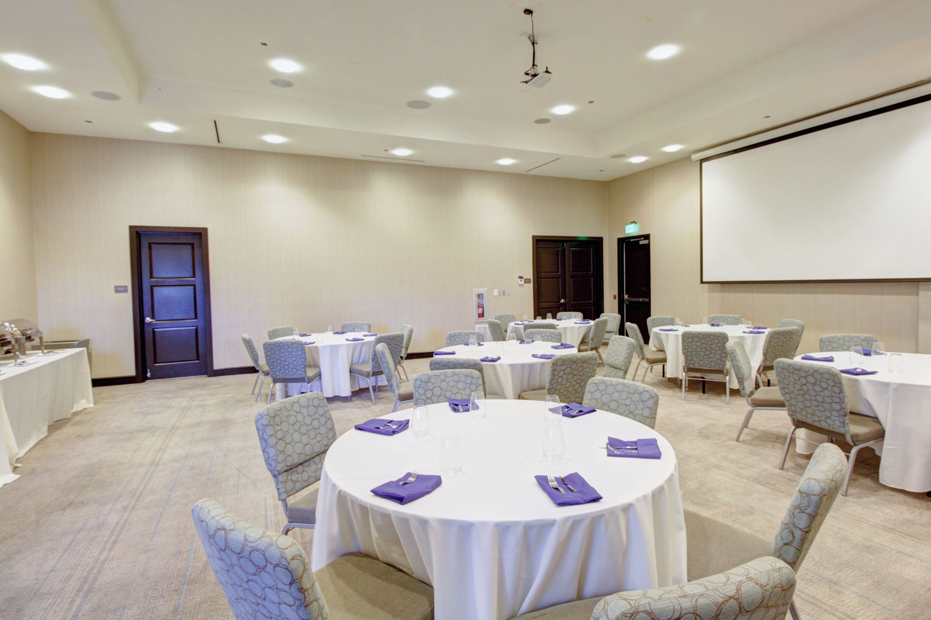 Un salón de conferencias con mesas redondas y sillas, una pantalla de proyector y una puerta.