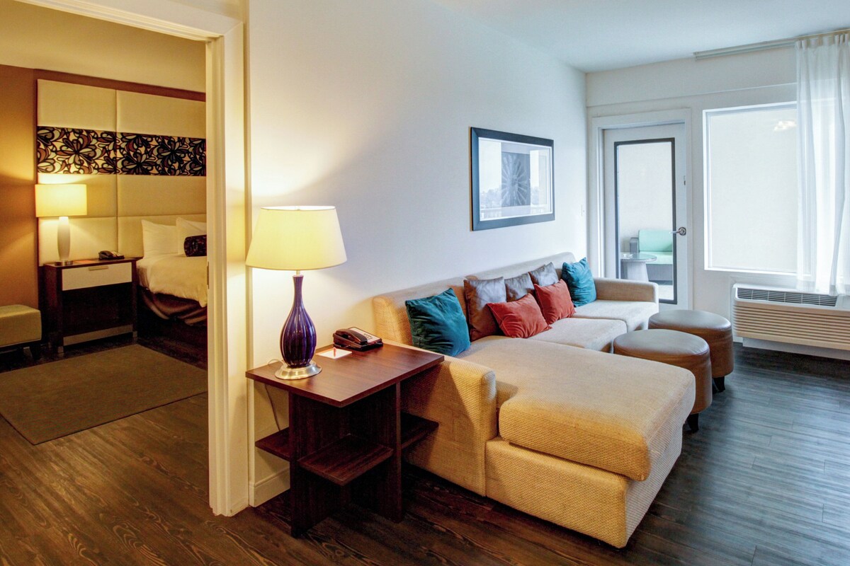 A hotel room with a sofa, a bedside table with a lamp, a chair, a bed, and a window with curtains.