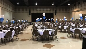 A large banquet hall with tables arranged in rows, a stage with balloons, and a projector screen.