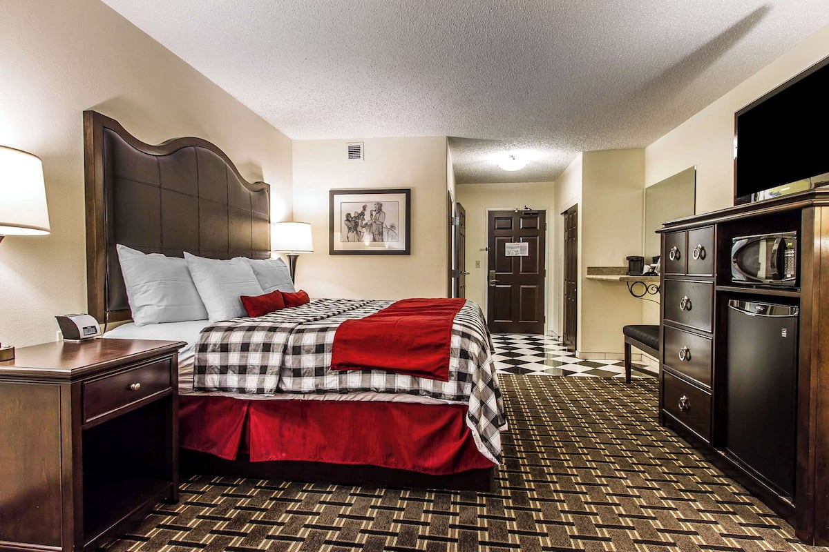 A hotel room with a large bed, a desk with a lamp, a dresser, a microwave, and a door leading to the bathroom.