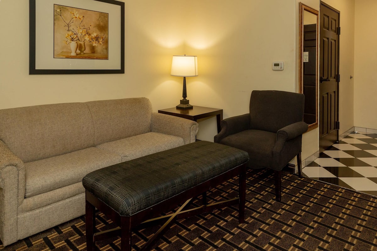 A room with a beige sofa, a dark armchair, a small table with a lamp, and a patterned carpet.