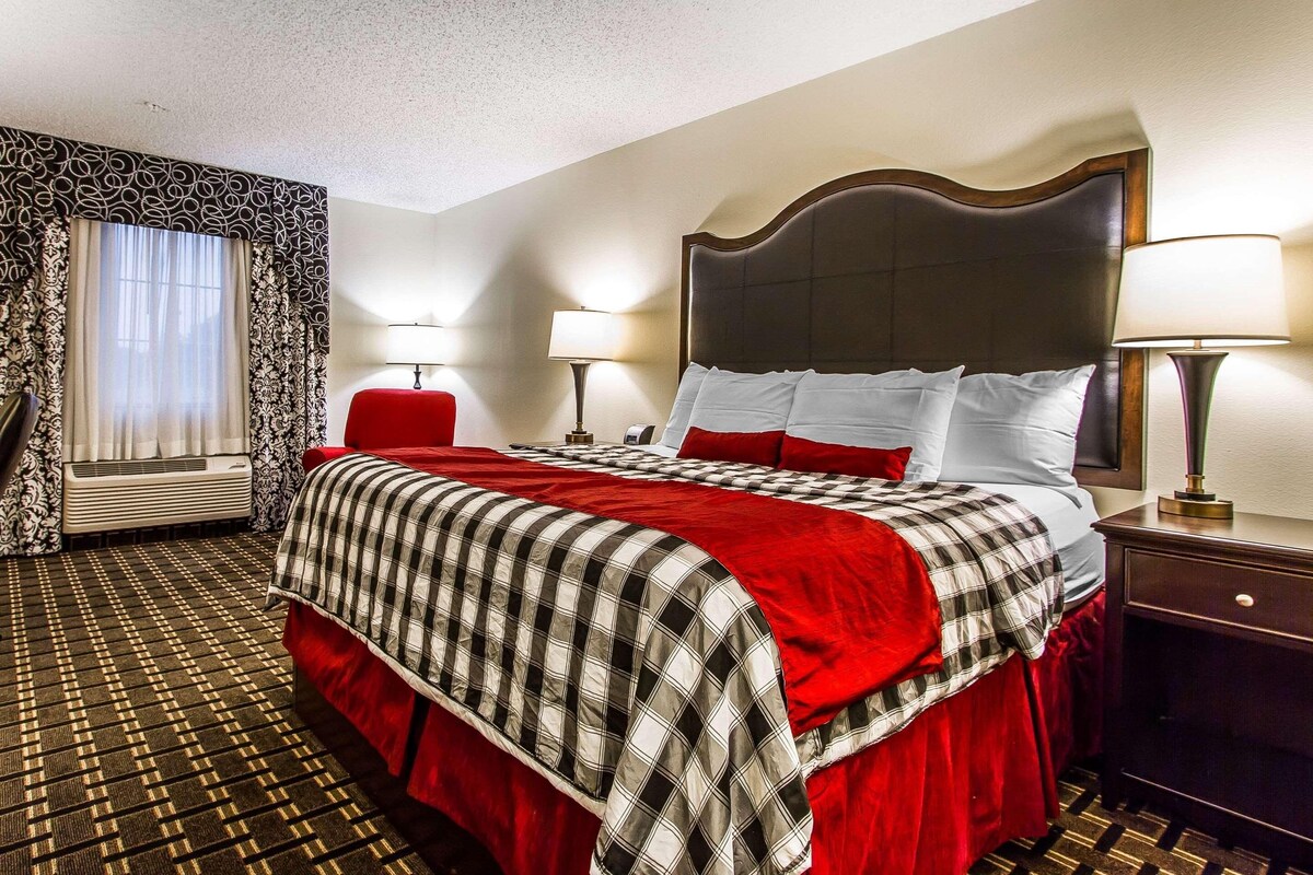 A hotel room with a large bed, two bedside lamps, a red chair, a window with curtains, and a patterned carpet.