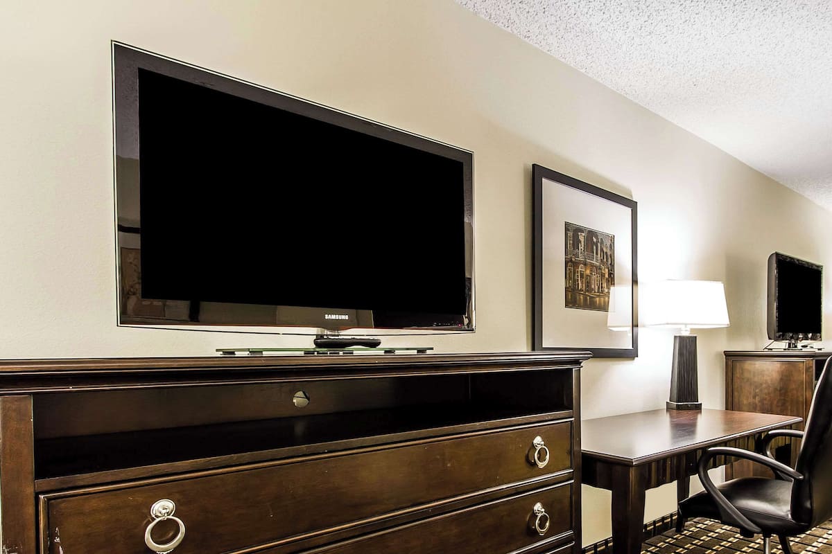 A hotel room with a large flat-screen TV mounted on the wall, a wooden dresser with drawers, a small table with a lamp, and a chair.