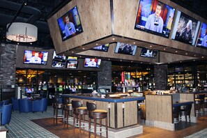 Zona de bar con varios televisores instalados en el techo, barra y distintas áreas de estar con taburetes.