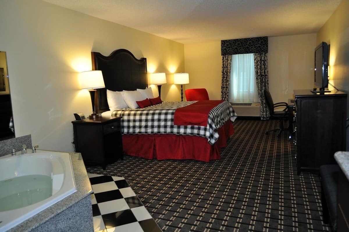 A hotel room with a checkered floor, a bed with a red and black bedspread, two bedside lamps, a nightstand, a large mirror, a television, a desk with a chair, and a window with curtains.