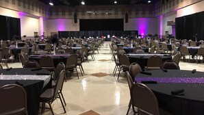 A large hall with rows of tables and chairs, a stage with a backdrop, and purple lighting.