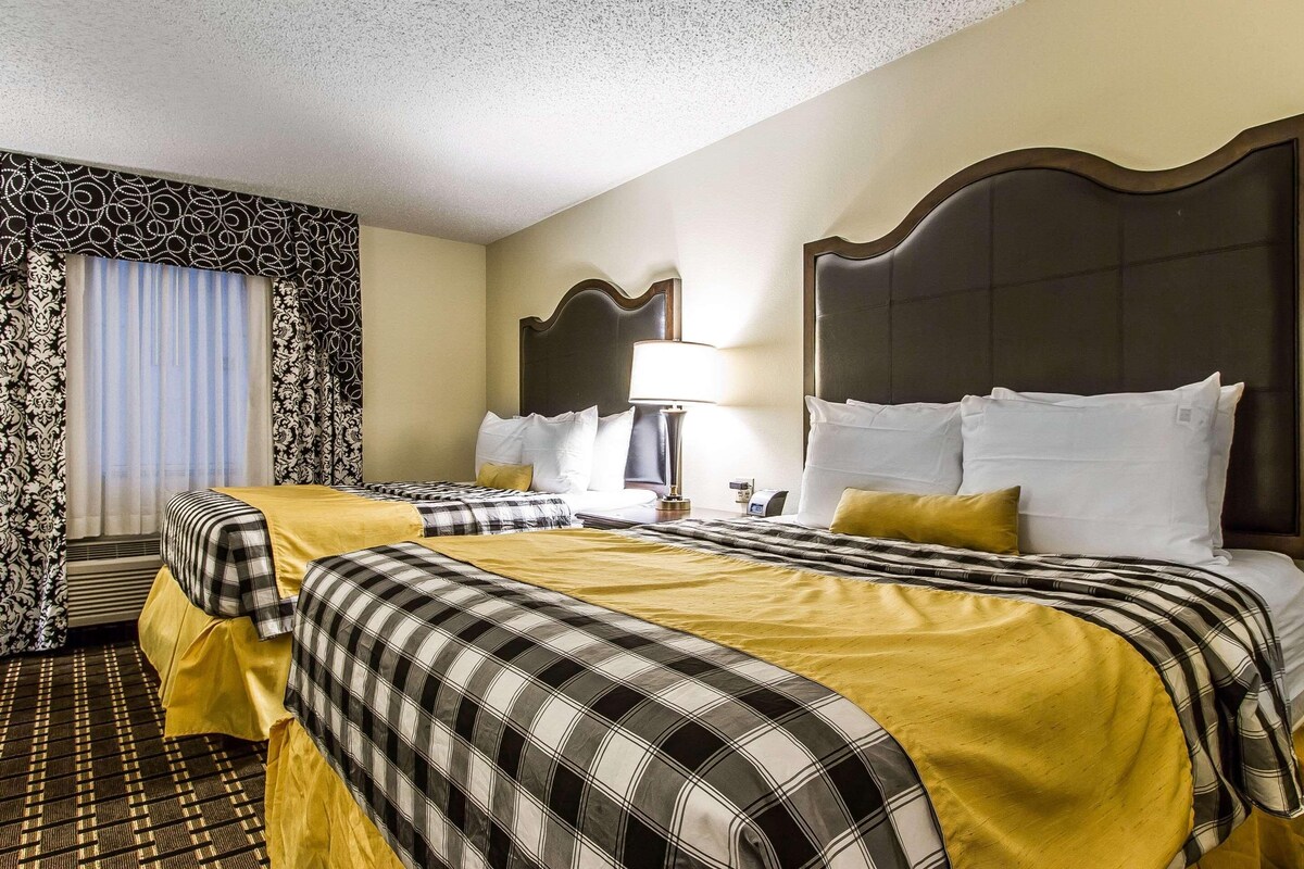 A hotel room with two beds, a large window with curtains, a headboard, and a bedside table with a lamp.