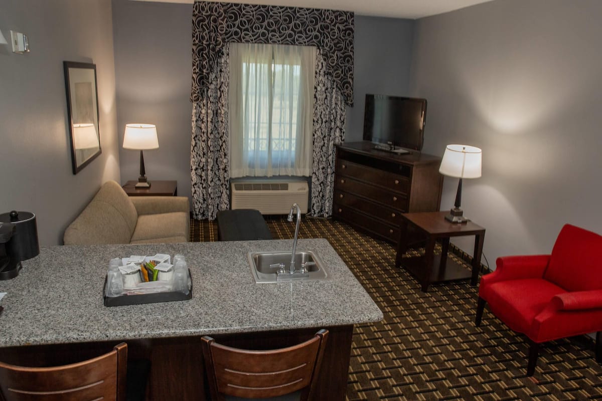 A hotel room with a kitchenette, a dining area, a red armchair, a television, and patterned curtains.