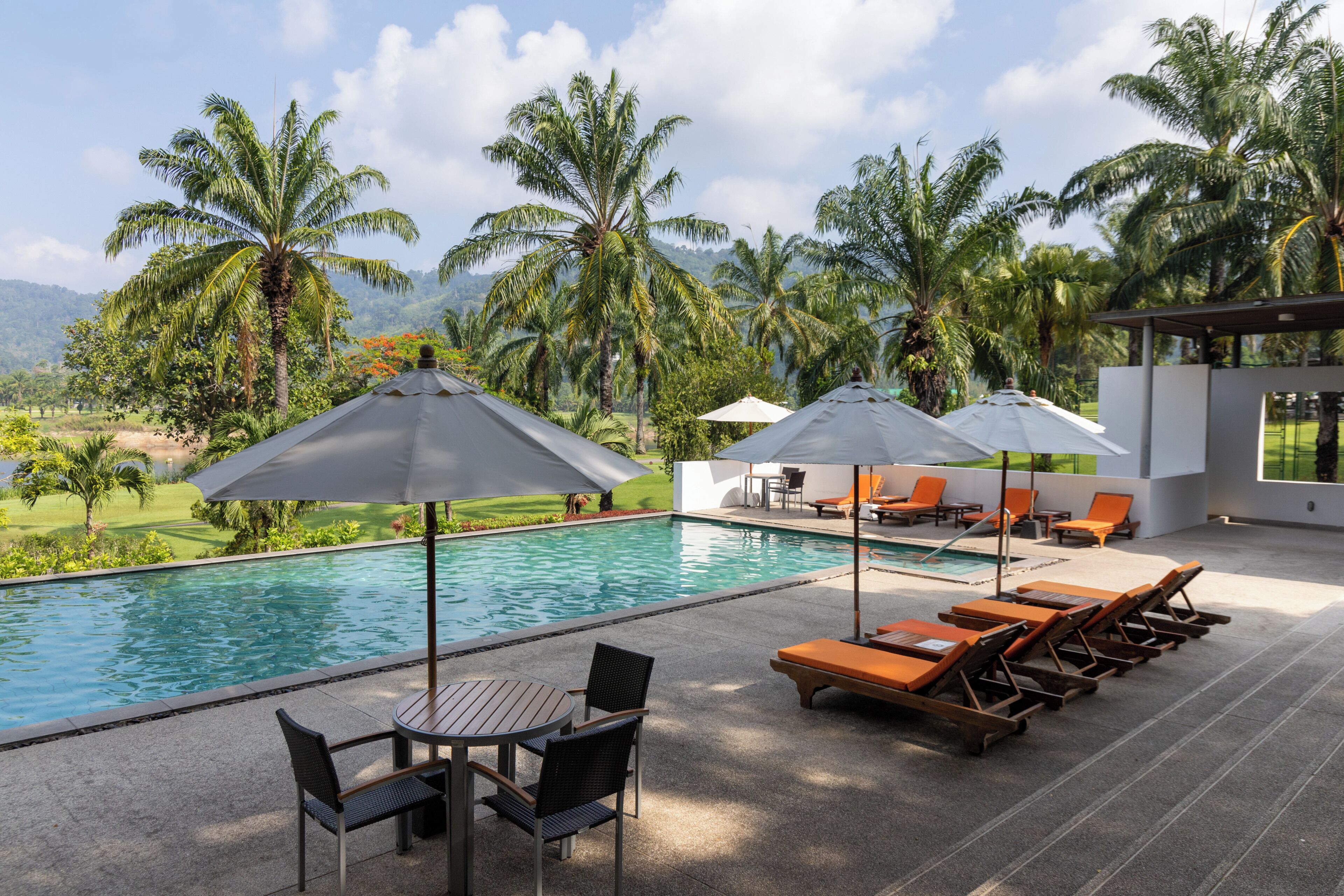 A pool area with lounge chairs and umbrellas, surrounded by palm trees and a view of a golf course.