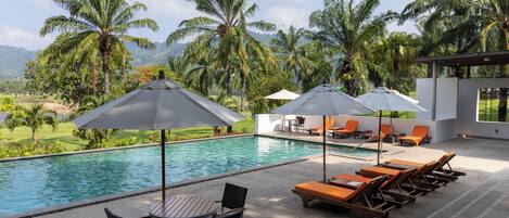 A pool area with lounge chairs and umbrellas, surrounded by palm trees and a view of a golf course.