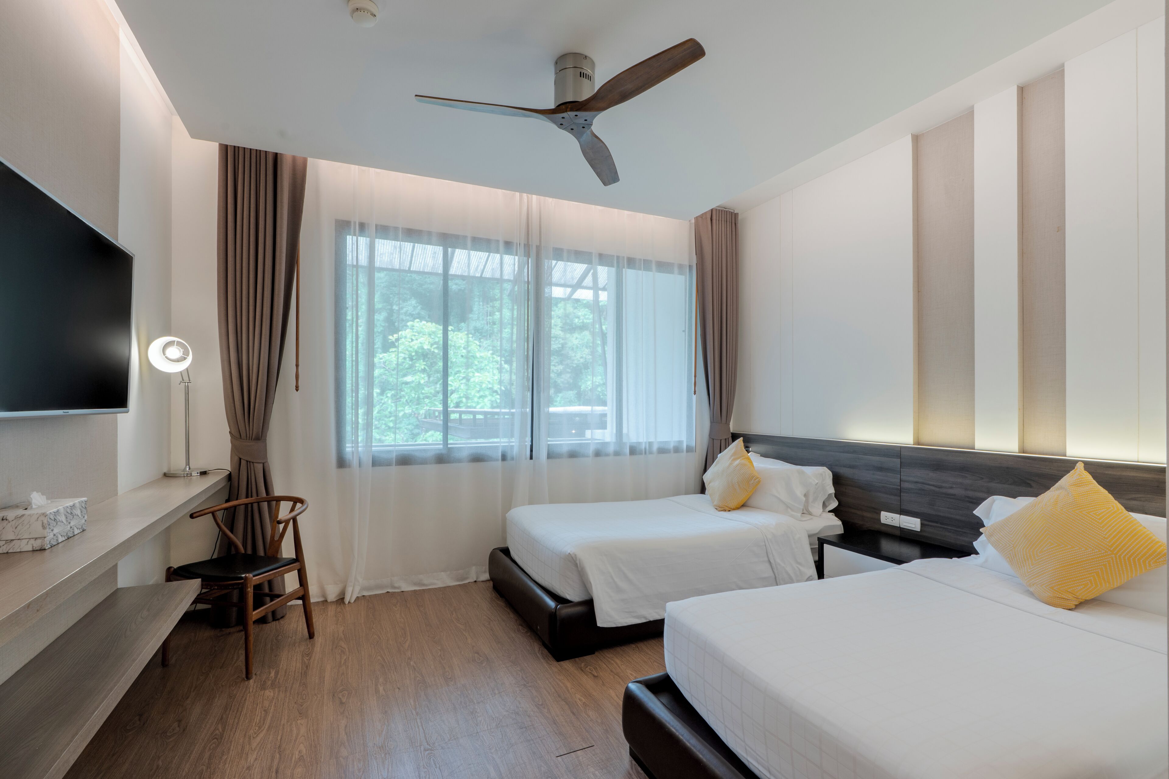 Habitación de hotel con dos camas, ventilador de techo, televisión, escritorio y una silla.