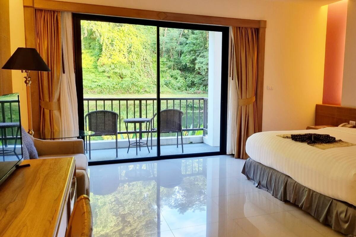 A hotel room with a large bed, a desk, a chair, and a balcony with a view of greenery.