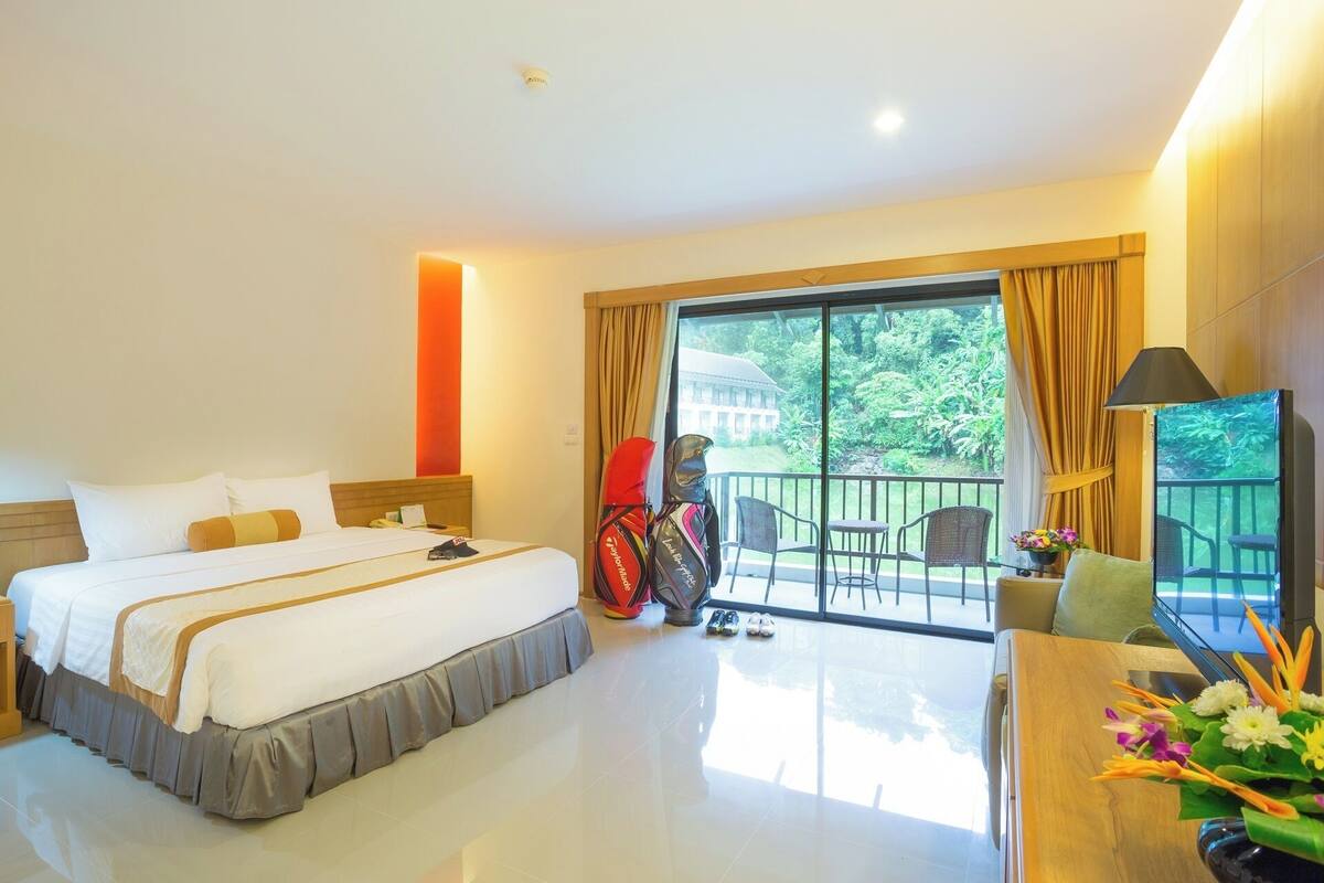 Habitación de hotel con cama, escritorio, silla, televisor y balcón con vistas a zonas verdes.