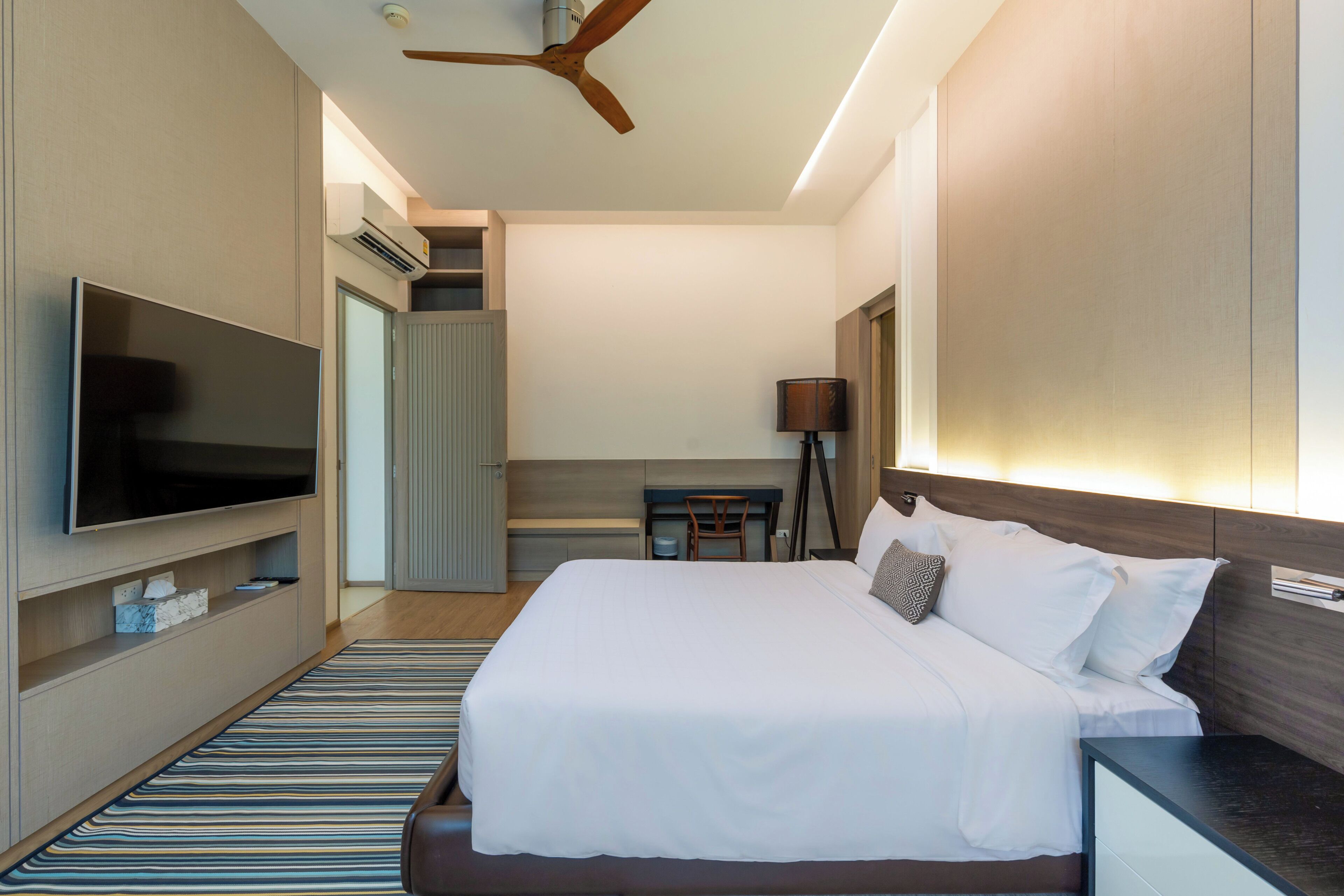 A hotel room with a bed, TV, ceiling fan, and a desk with a lamp.