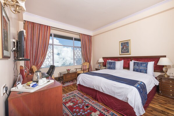 A hotel room with a large bed, a desk with a coffee maker, a chair, a window with curtains, and a painting on the wall.